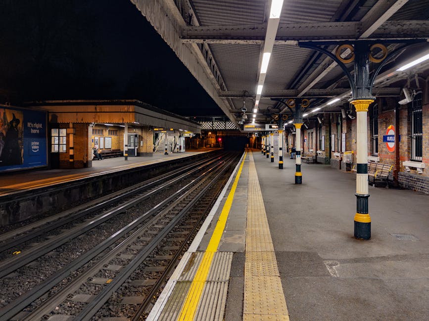 Inside a large railway station with a curved glass and metal arched ceiling, multiple train tracks extend into the distance, with platform edges visible on either side. A black station clock and digit