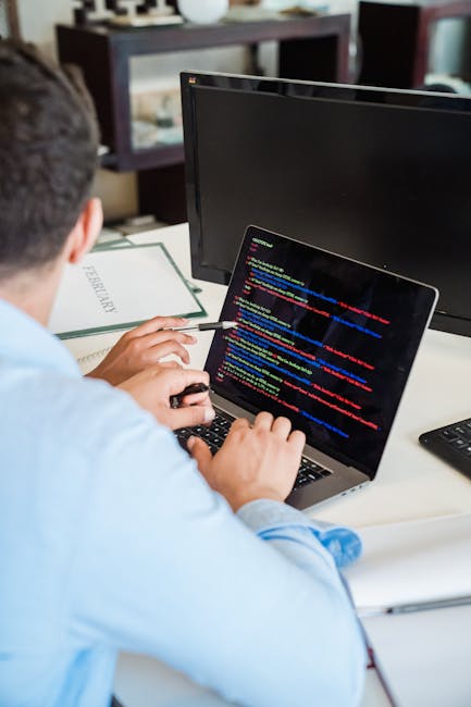 A person with short dark hair, dressed in a light blue shirt, seated at a desk during a home relocation or moving project, viewing a laptop with a screen displaying colorful lines of code or computer 